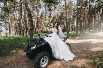 A beautiful bride in a white long dress sits on a black ATV in the forest. Wedding photography.