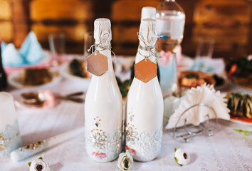 A white bottle of champagne, decorated with precious stones, jewelry with a metal, gold tag tied with a thread stands on a wooden table. Wedding photography.