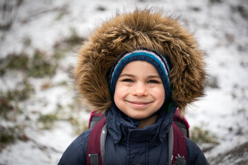 Portrait of smiling preschool boy in warm coat with hood in winter against snow