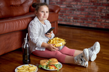 Fat girl eating chips from bowl while sitting on floor in living room, side view. Overweight...