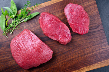 Raw dry aged bison beef rump steak piece and slices offered with herbs as top view on wooden design...