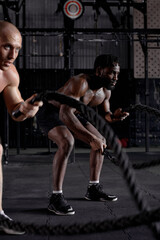 Fototapeta na wymiar two diverse athletic guys in dark cross fit class, males started training exercises get sweating, holding battle ropes in hands, concentrated and motivated. sportive active healthy lifestyle.
