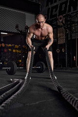 Muscular caucasian man working out with heavy ropes. handsome man with perfect body in dark sports club gym. Strength and motivation. Active intense cross fit training, male full of energy