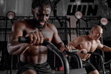 Fototapeta na wymiar Men exercise bike gym cycling training fitness. Two Fitness male using air bike cardio workout. Two guys biking indoor at gym, exercising legs. Cross functional training. focus on black male