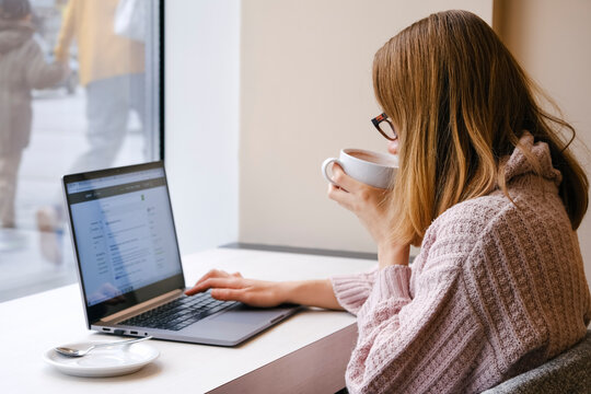 Young attractive woman working on laptop in modern coworking space and drinking cocoa. Elerning or freelance concept.