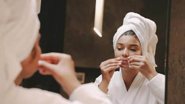 A young dark-skinned brunette in front of a mirror in a dressing gown and with a towel on her head uses a patch for lips, the girl is reflected in the mirror. skin care. High quality 4k footage