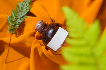 Fototapeta premium 50 ml Perfume bottle with blank white label and green leaves on textured orange fabric background. Perfume bottle stock photo