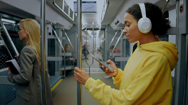 A young girl standing in modern metro train, wearing headphones and looking in her phone. A man walks past her, obscuring the frame. She is wearing a bright yellow hoodie and white headphones. 