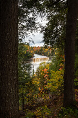 Beautiful autumn photograph of Upper Tahquamenon Falls in Michigan with water cascading into the river below framed between trees with fall colored foliage.