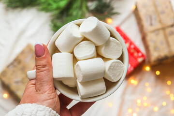 Woman's hand holding a cup with marshmallows on blurred holiday background. Winter, festive and...