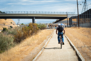 Photo of a young man in a baseball cap and backpack biking along the American River Parkway trail...