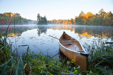 Yellow canoe on shore of calm lake with island at sunrise during autumn - obrazy, fototapety, plakaty