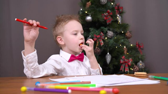Blond little boy with bow tie holding red marker eats tasty candy sitting AT table with papers by decorated Christmas tree at home