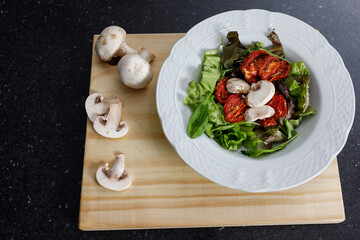 Close up dish with lettuce salad, arugula, sun-dried tomatoes and white mushrooms. On wooden board with whole mushrooms. Vegetarian or vegan food. Copy space for text.