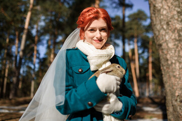 beautiful girl in a green coat and a wedding dress holds a little rabbit in her arms