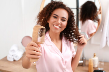 Young African-American woman with wooden comb in bathroom - obrazy, fototapety, plakaty
