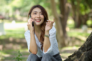 Fototapeta premium Happy female talking on the phone, sits under the tree in the park