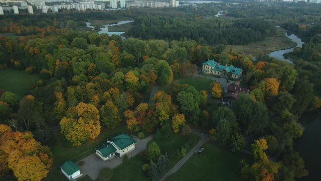 Flight over the autumn park. Trees with yellow autumn leaves are visible. Park buildings are visible. On the horizon there is a blue sky with clouds and city houses. Aerial photography.