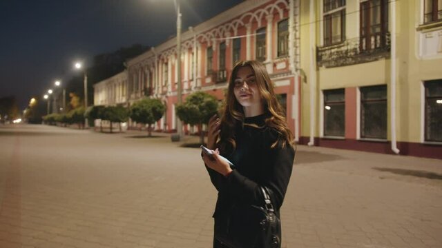 A spectacular young girl goes in the evening through the historic quarter of the city and leafs through the phone screen. Side view