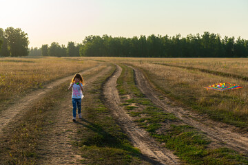 Fototapeta na wymiar Little girl with rainbow colored kite running along the country road in the summer field. Selective focus.