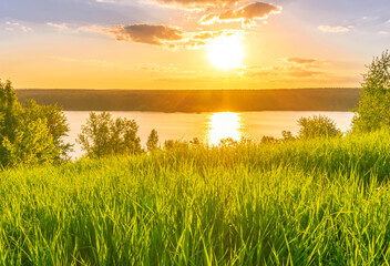 beautiful sunrise or sunset view from grass to a moutain lake with sun glow and cloudy sky above the water and forest on the background