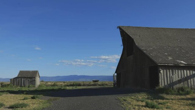 Static footage of a large and a small barn standing still on a blue sunny day with fields and mountains in the background. Sunny day, swifts fly into their nests.