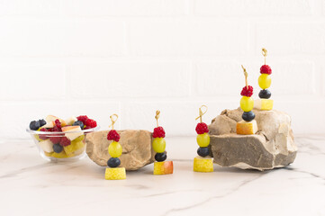 canapes of juicy fruit on skewers, a mix of cut fruit in a glass bowl. composition on stones and marble table. front view.