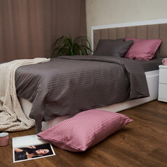 A large bed with a brown-gray bedding(linens) and pink pillows are in the bedroom. There is also a white blanket, scattered pillows, a magazine and a cup on the floor.