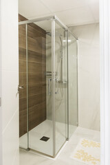 Fragment of bathroom interior with tempered safety glass enclosure with shower fittings head hanging on tiled wall. Modern minimalist style practical design materials