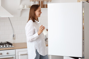 Profile portrait of attractive dark haired woman wearing white shirt, looking smiling inside fridge...