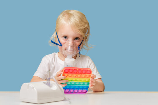 A happy child in therapeutic inhalation mask is sitting at a table and playing pop it toy. Treatment of colds at home. Copyspace.