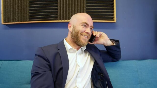 Bald businessman laughing and alking With A Friend On Mobile cell Phone While Stitting On Sofa in Home Office
