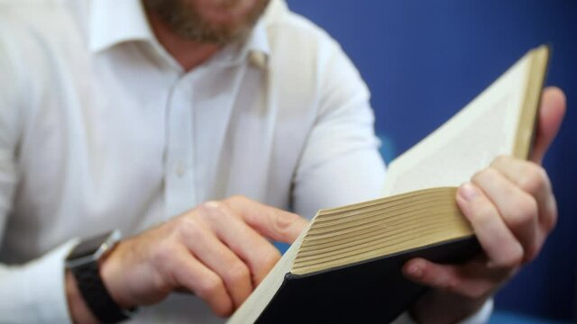 A man holding and Reading A Book with Fingers following The Lines. The Unrecognizable male is wearing a white shirt.