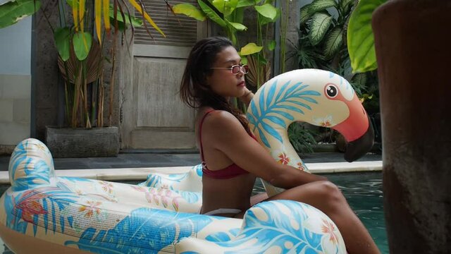 Asian bikini model sitting on floating rubber swan swimmingpool toy enjoying her best life at a private pool. Close up slide shot following the subject. Holiday in Bali. Lush green villa garden.