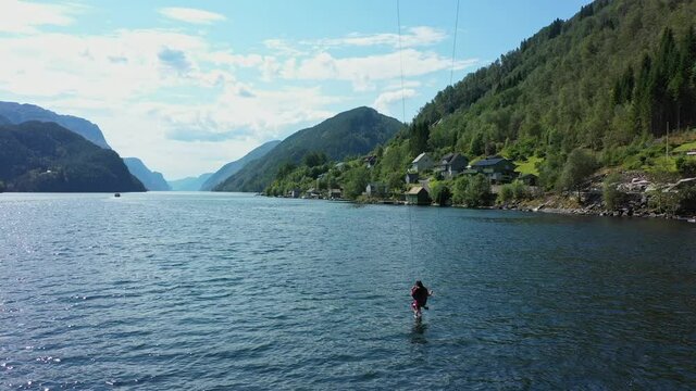 Woman riding spectacular tall swing with Norwegian fjord Veafjord in background - Aerial showing woman dangling her feet down into sea surface surrounded by stunning scenery - Norway
