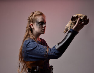 Closeup portrait of young red haired woman with black barbarian facepaint wearing medieval viking inspired costume Holding  sheep skull for ritual  posing against studio background with red lighting