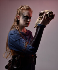 Closeup portrait of young red haired woman with black barbarian facepaint wearing medieval viking inspired costume Holding  sheep skull for ritual  posing against studio background with red lighting
