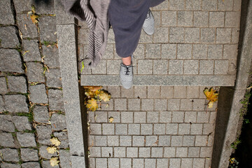 Fragment of a woman's leg in navy blue pants and white and gray shoes with a geometric pattern on a cobblestone staircase with lying yellowed leaves, top view.