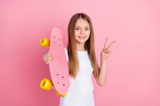 Photo of funny blond little girl show v-sign hold skate wear white t-shirt isolated on pink color background