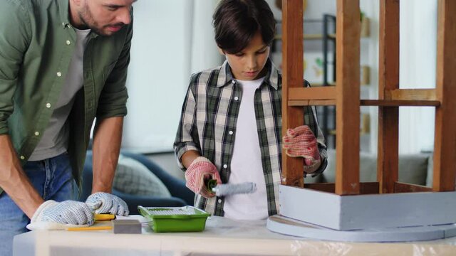 renovation, diy and home improvement concept - father and son in gloves with paint roller painting old wooden table in grey color at home