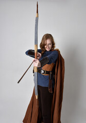 
Full length, portrait of red haired woman wearing medieval viking inspired costume and flowing cloak,  Holding bow an arrow weapon,  posing against studio background.