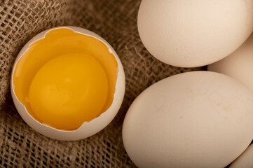 A broken white chicken egg and several whole eggs on a homespun fabric with a rough texture, close-up selective focus.