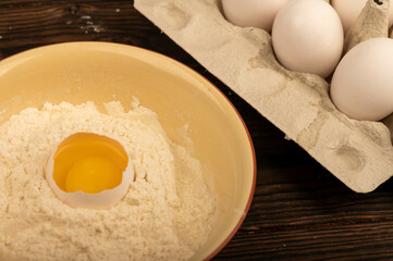A broken white chicken egg in a bowl of wheat flour and several whole eggs in a tray made of white cardboard, close-up selective focus.