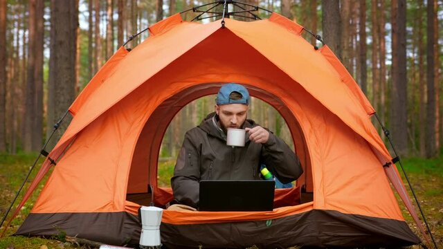 Young male freelance tourist remotely works at computer, sitting in an orange tent camping in nature in forest, drinking hot coffee and working on laptop. Working startup project at laptop in nature.