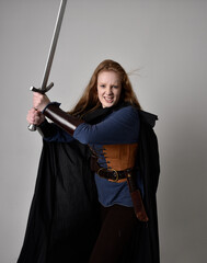 Close up portrait of red haired woman wearing medieval viking inspired costume and flowing cloak,  Holding a long sword weapon, action pose against studio background.
