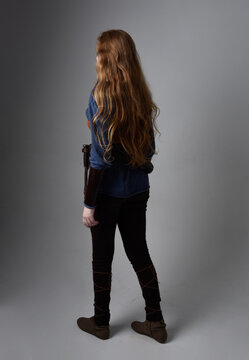 Full Length Portrait Of Red Haired Woman Wearing Medieval Viking Inspired Costume,  Standing Pose With Back To The Camera, Against Studio Background.