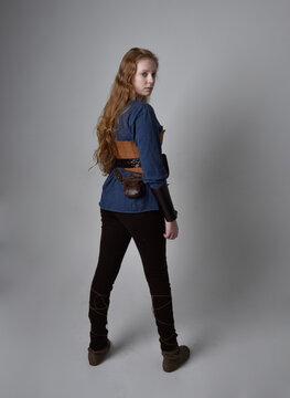 Full Length Portrait Of Red Haired Woman Wearing Medieval Viking Inspired Costume,  Standing Pose With Back To The Camera, Against Studio Background.