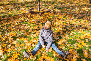 A teenager sits on autumn leaves in a park with headphones on and listens to music with his eyes closed.