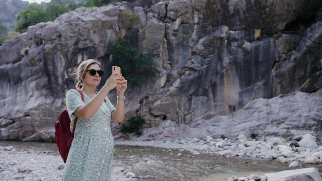 a pregnant girl in dress with a backpack stands near a mountain river and rocks, and takes pictures on the phone. Beautiful landscape. The wind develops her dress. Travel, vacation.