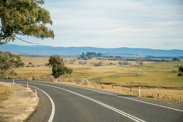 Open empty road surrounded by farms and fields in Australia. Mountains on the horizon. Road trip travel